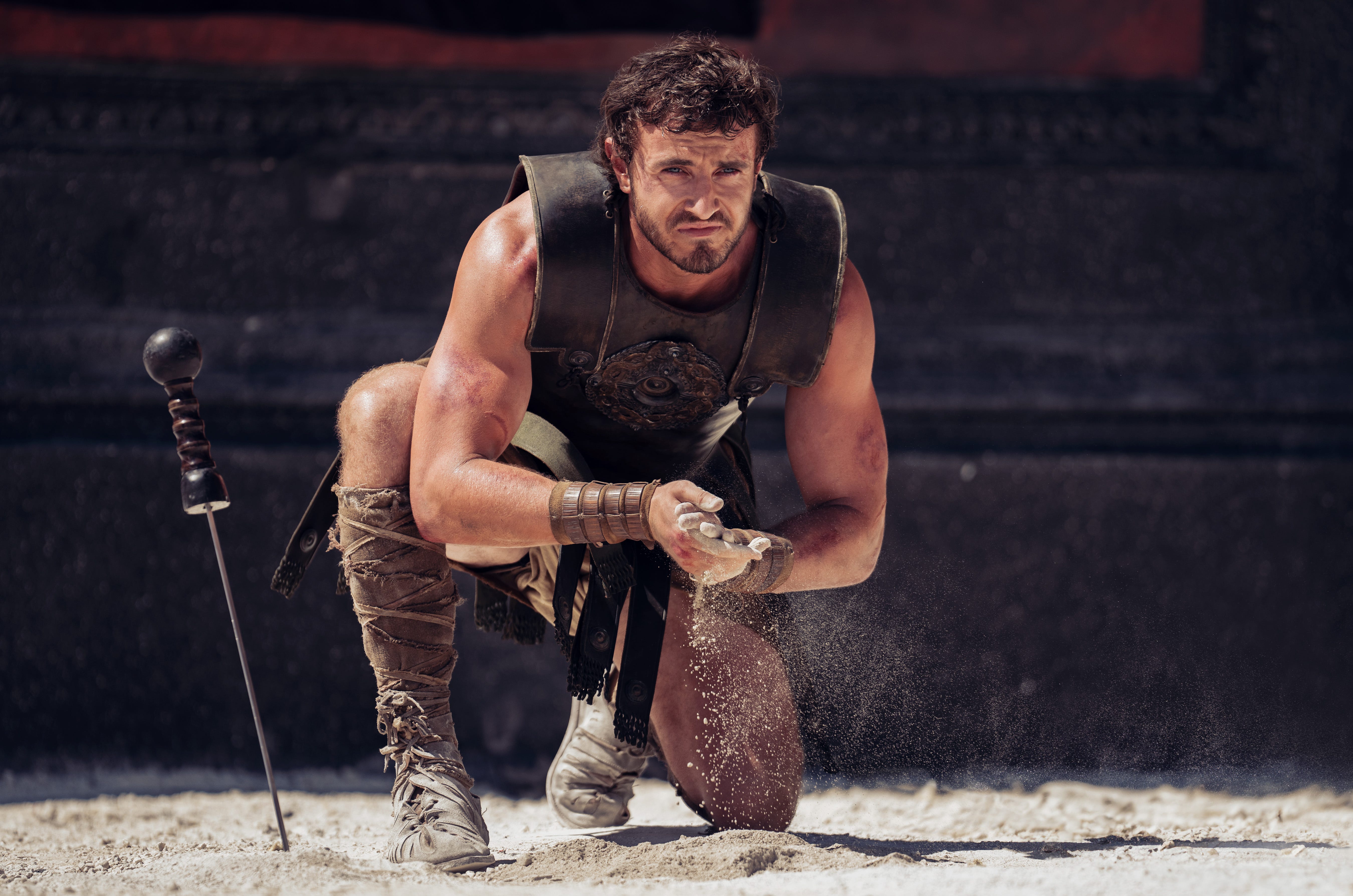 paul mescal as lucius in gladiator two. he's crouching on the dirt floor of the colosseum, his sword stuck in the ground, as he rubs dirt between his hands