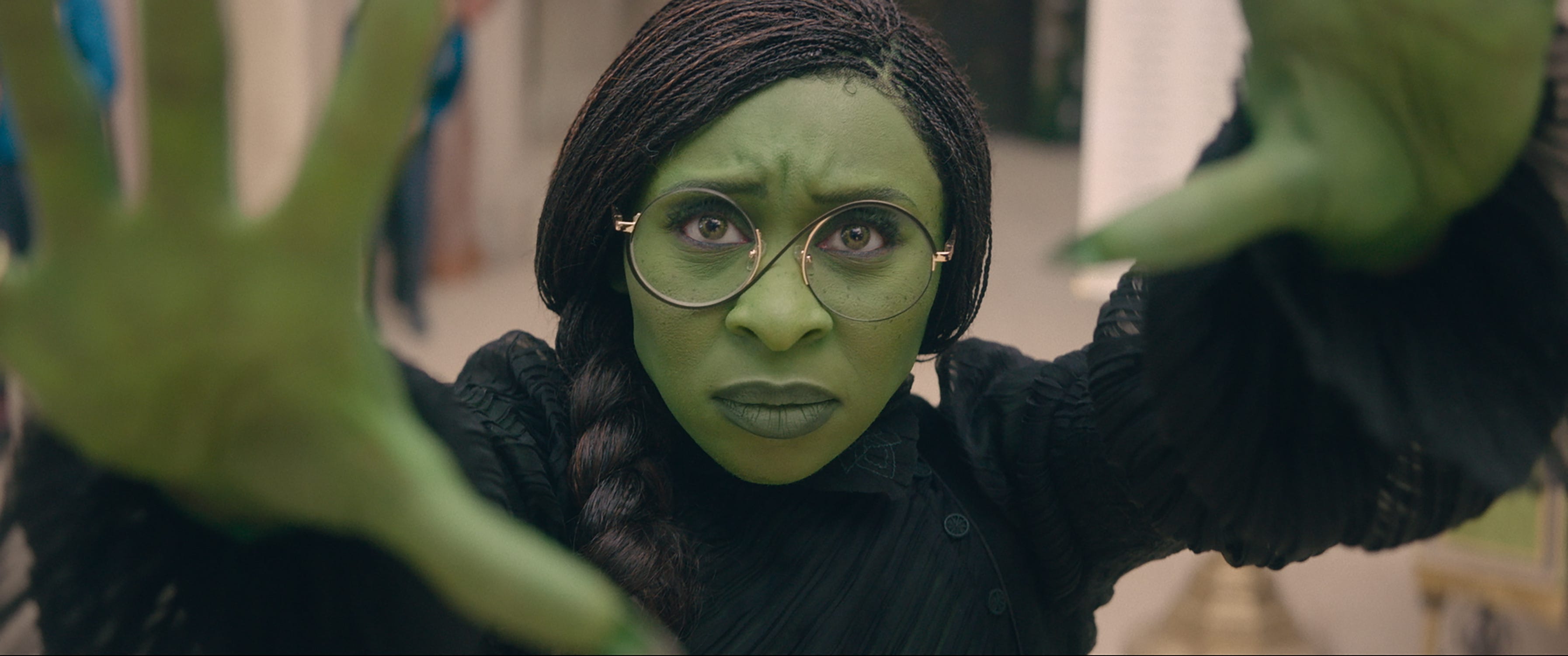 cynthia erivo as elphaba in wicked. she's painted green and is wearing a black dress, with her hair styled in microbraids braided into one larger braid over her right shoulder. her hands are outstretched, and her expression is intent