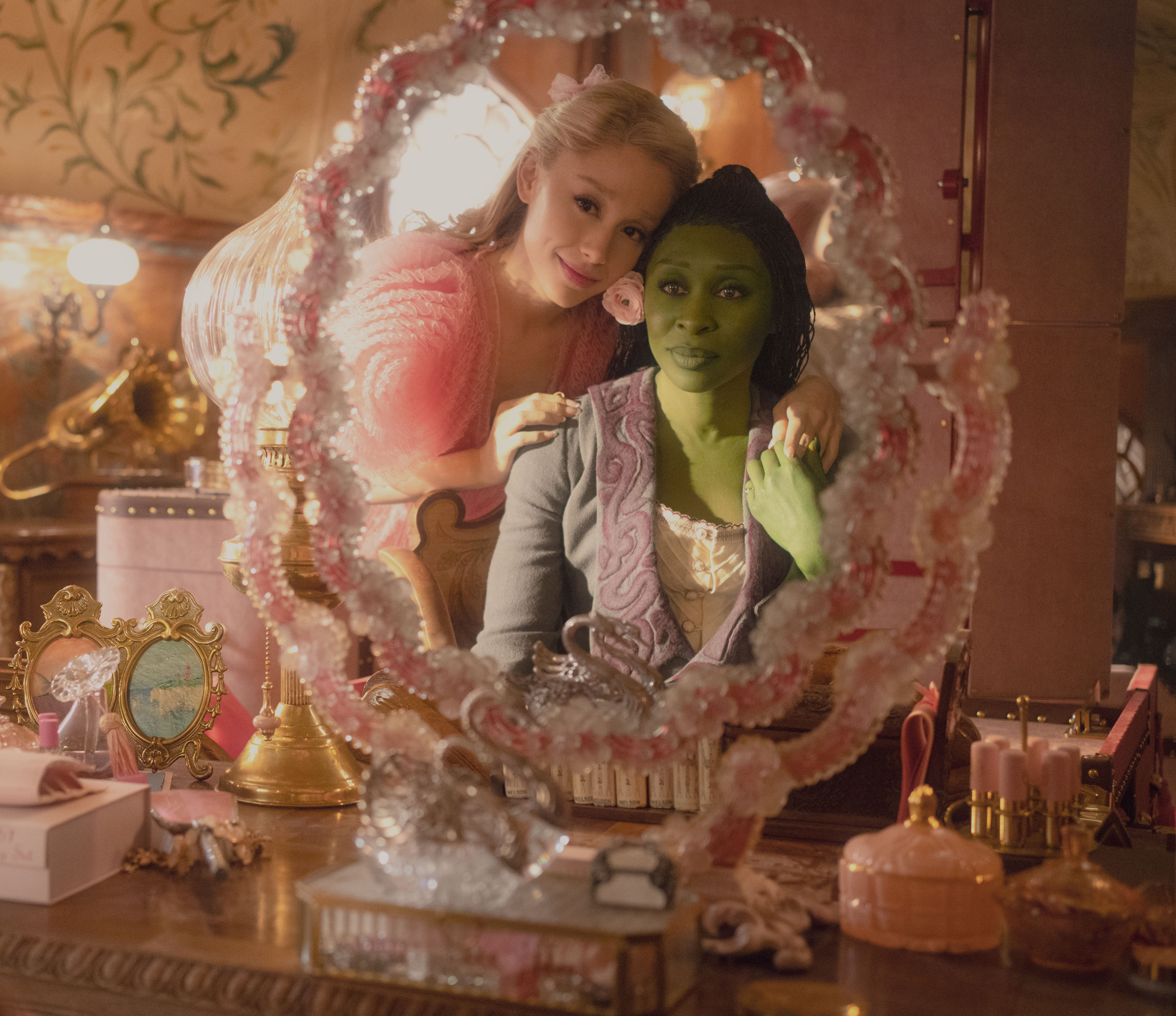 A still from "Wicked" shows Ariana Grande wearing a pink dress and Cynthia Erivo, painted green, wearing a purple robe. They are reflected in a mirror on a table.