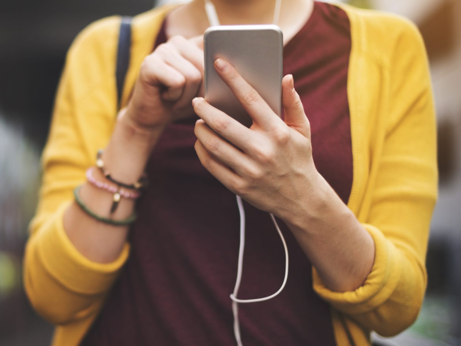 A close-up of a woman wearing a dark-red shirt and a yellow sweater holding a phone and wearing headphones. Her face is not visible.