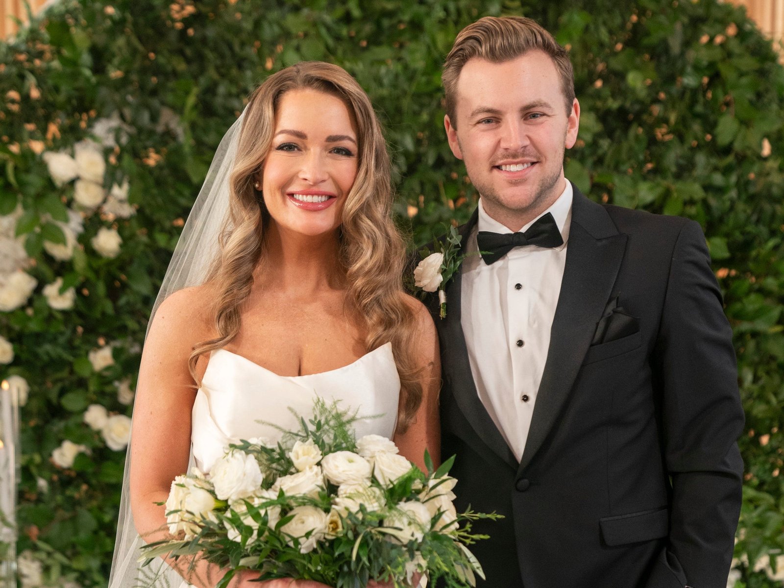 A still of "Love Is Blind" showing Taylor in a wedding dress, holding a bouquet of white roses, standing next to Daniel in a tuxedo.