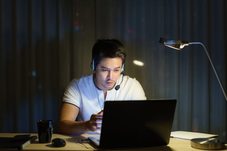 Man working from home at night on his laptop, wearing a headset.