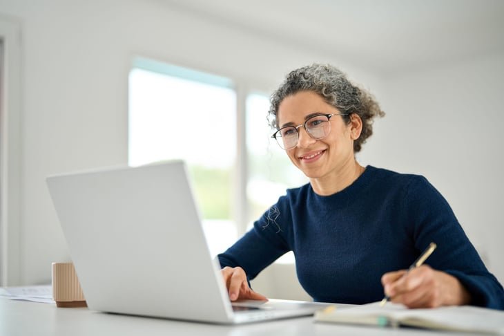 Happy middle-aged woman sitting at table at home office using laptop to work remotely