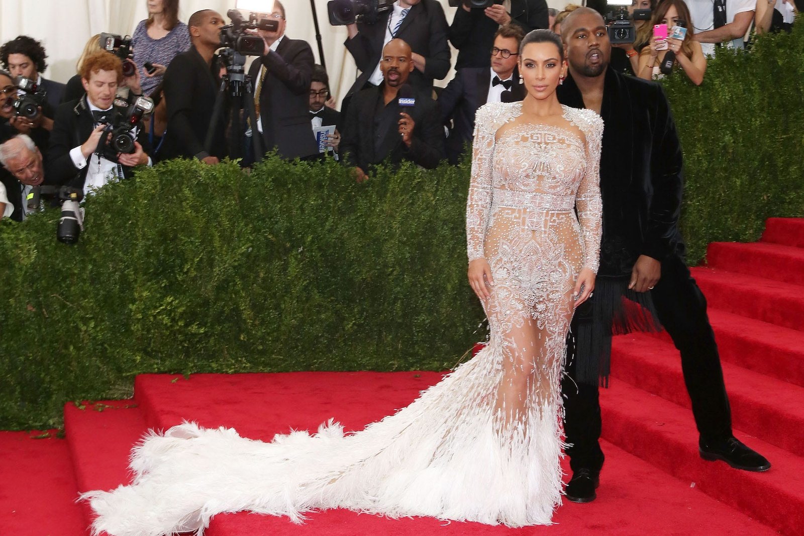 Kim Kardashian poses for photos with then-husband Kanye West on the Met Gala red carpet. She's wearing a nude gown with silver embellishments and a white feathered skirt while he's wearing a black suit with a velvet jacket.