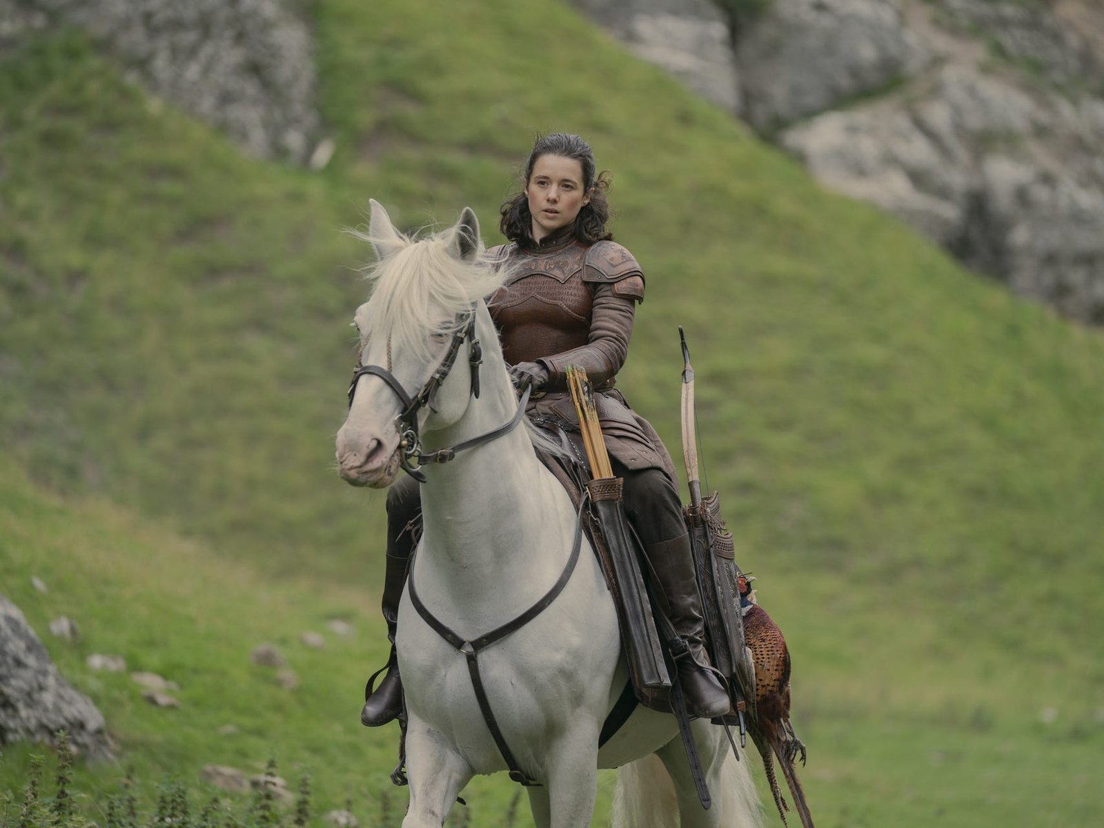 rachel redford as lady rhea royce in house of the dragon, a young woman with brown hair pulled half back, wearing brown armor and sitting astride a white horse, riding through a grassland
