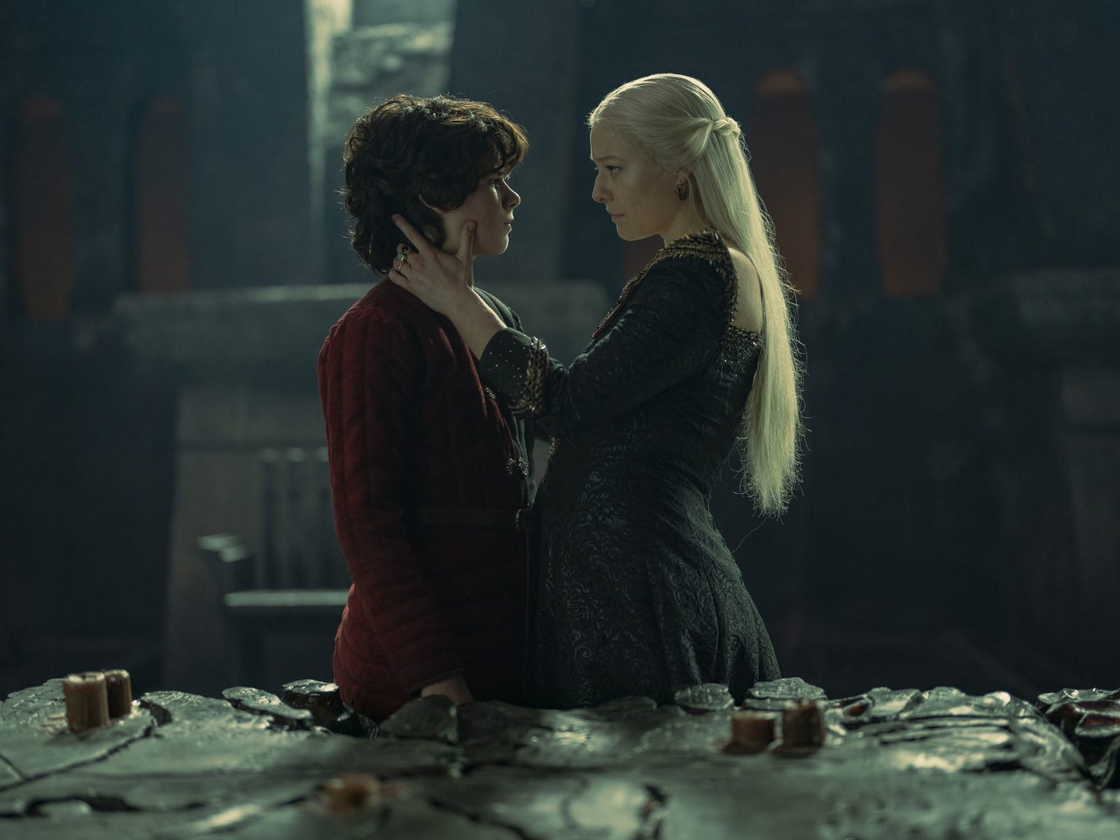 rhaenyra, emma d'arcy, holding her sun lucerys, elliot grihault, in front of a strategy war table. she's looking at him intensely, and cracling his neck