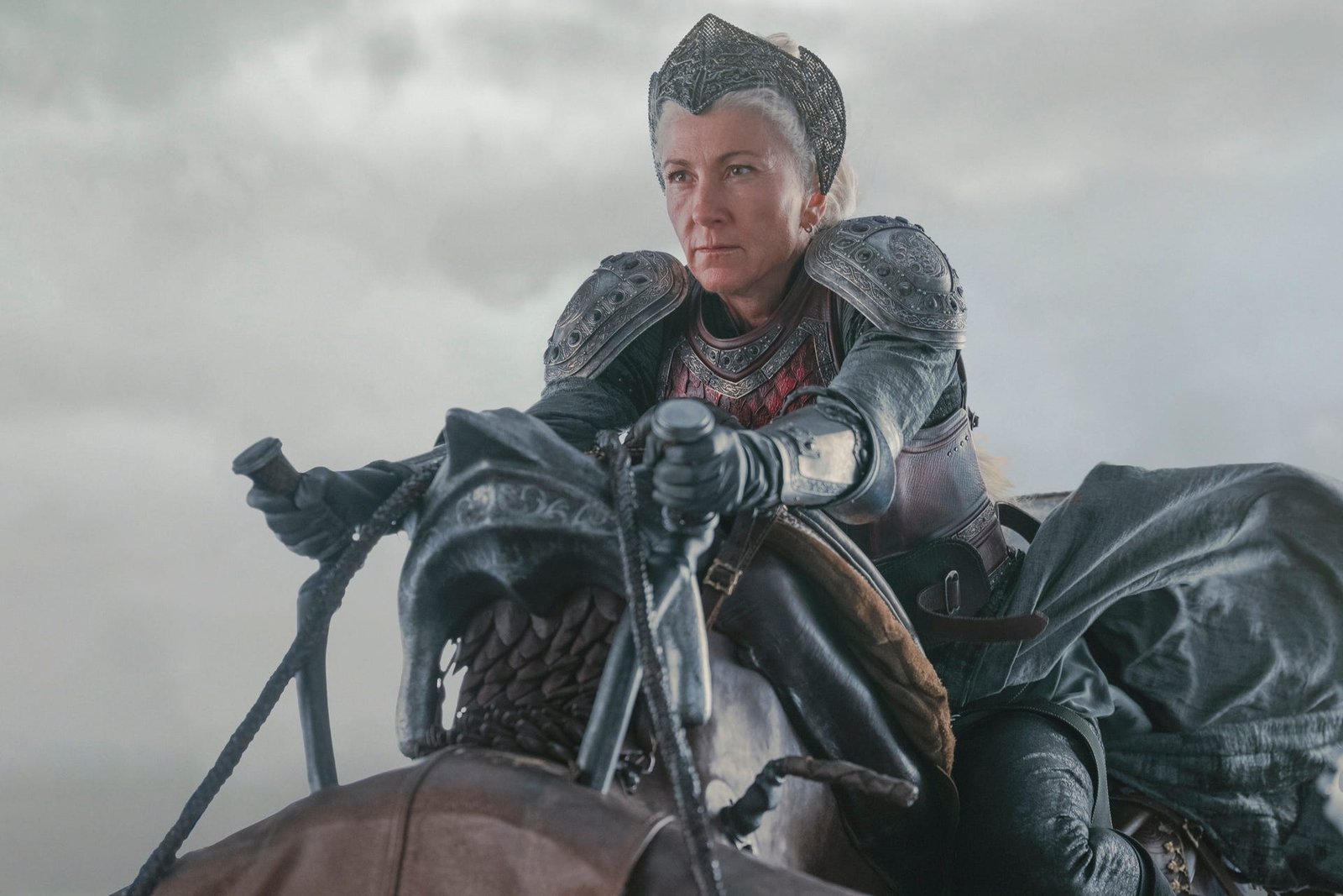 eve best as rhaenys targaryen in house of the dragon, dressed in full armor and a headpiece while clinging onto a dragon statue. she's flying through a grey sky, and her expression is resolute
