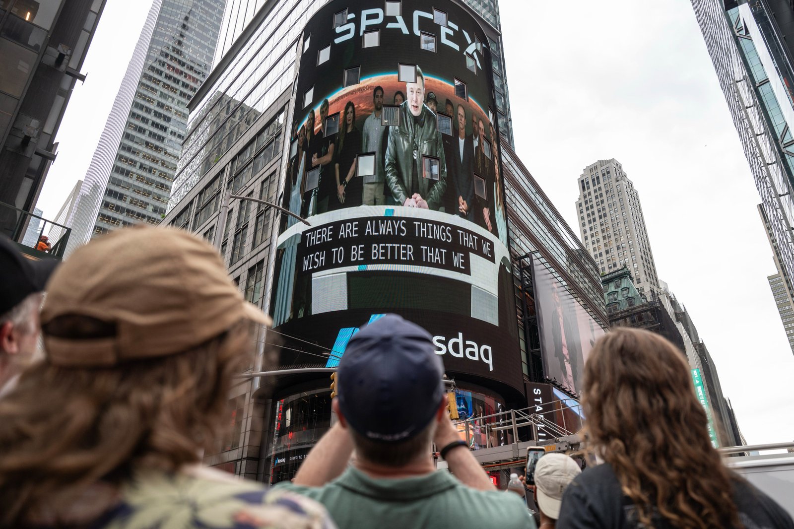 Crowd watches a Times Square Nasdaq billboard displaying SpaceX footage with Elon Musk and caption text.